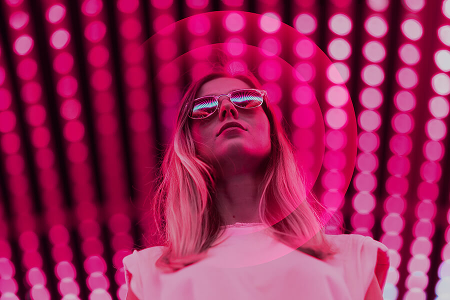 A women wearing reflective sunglasses stands boldy infront of pink neon lights