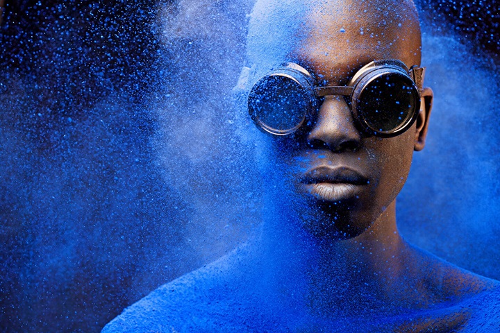 A person wearing round, steampunk-style goggles stands covered in vibrant blue powder, with more powder exploding across their face and shoulders. The background is dark, making the swirling blue particles stand out dramatically. The person’s expression is calm and neutral as the pigment envelops them.