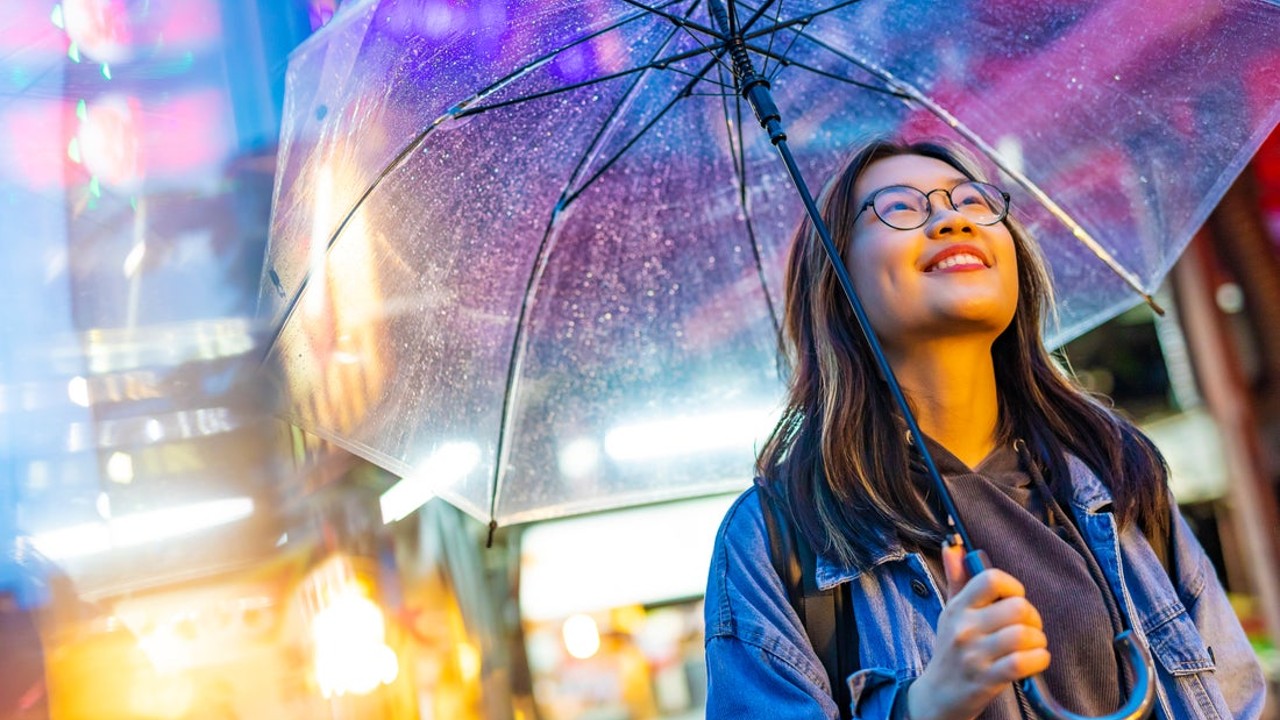 Smiling young woman with glasses holding a clear umbrella at night, illuminated by colorful city lights and neon signs.